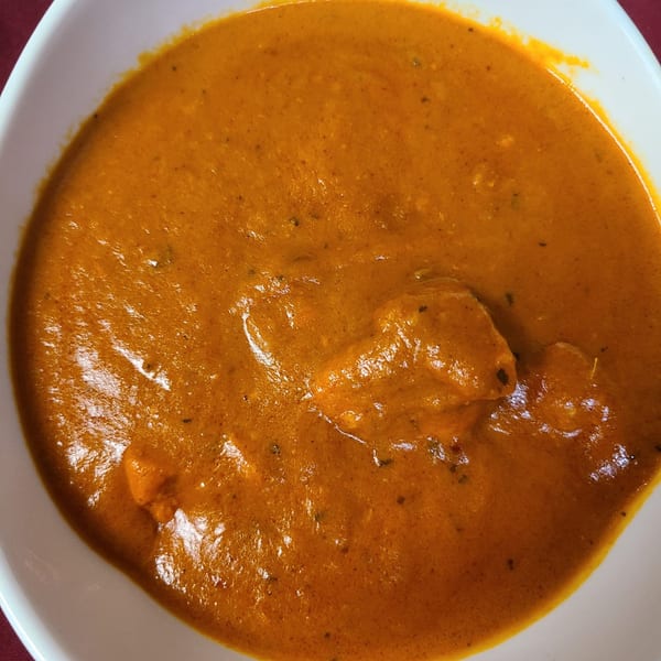 Close-up of a white bowl filled with vibrant orange Indian curry, featuring chunks of chicken.